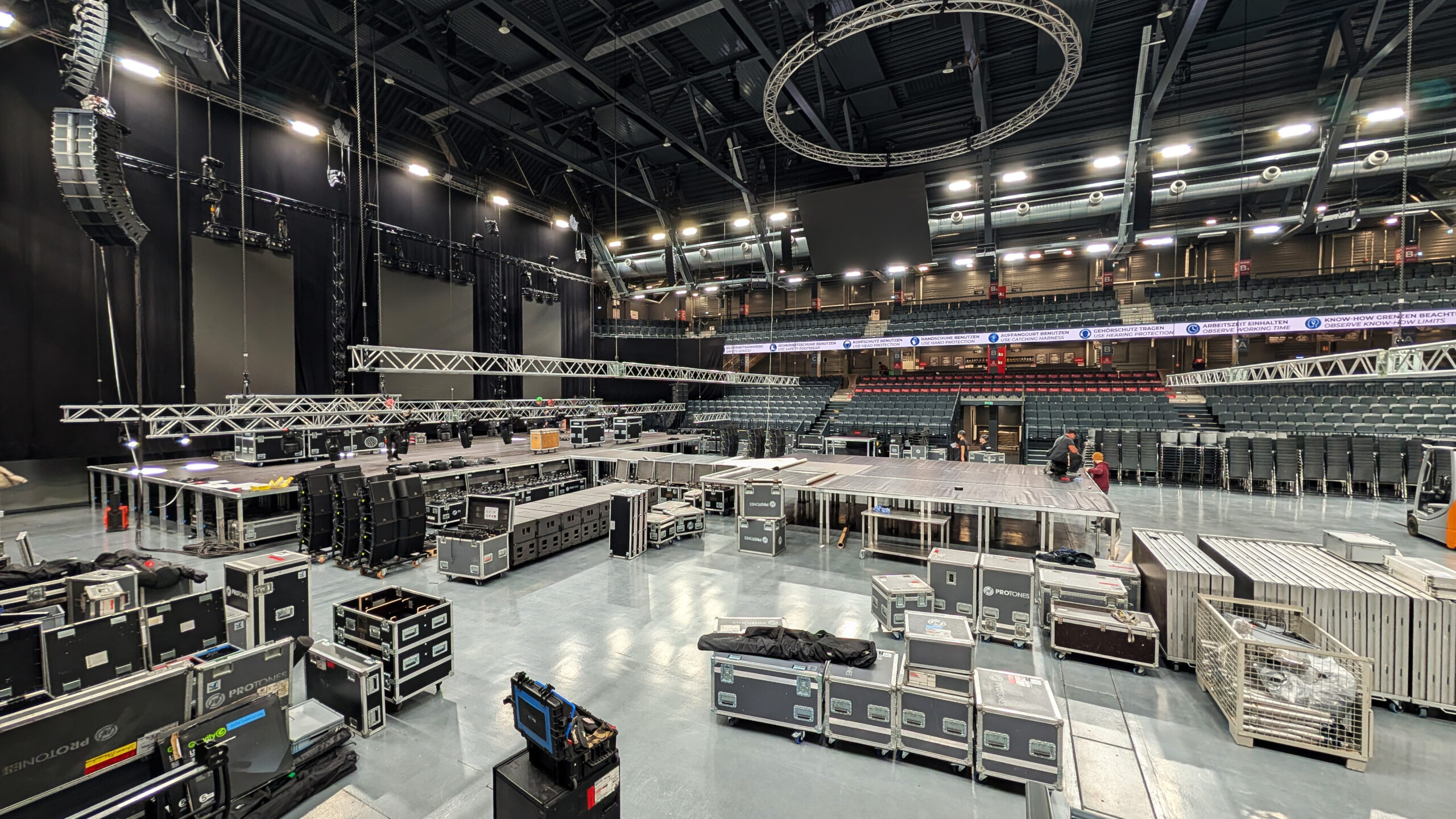 Innenansicht einer großen, leeren Halle mit aufgebauten Bühnenelementen, Traversen, Lautsprechern und Flightcases.