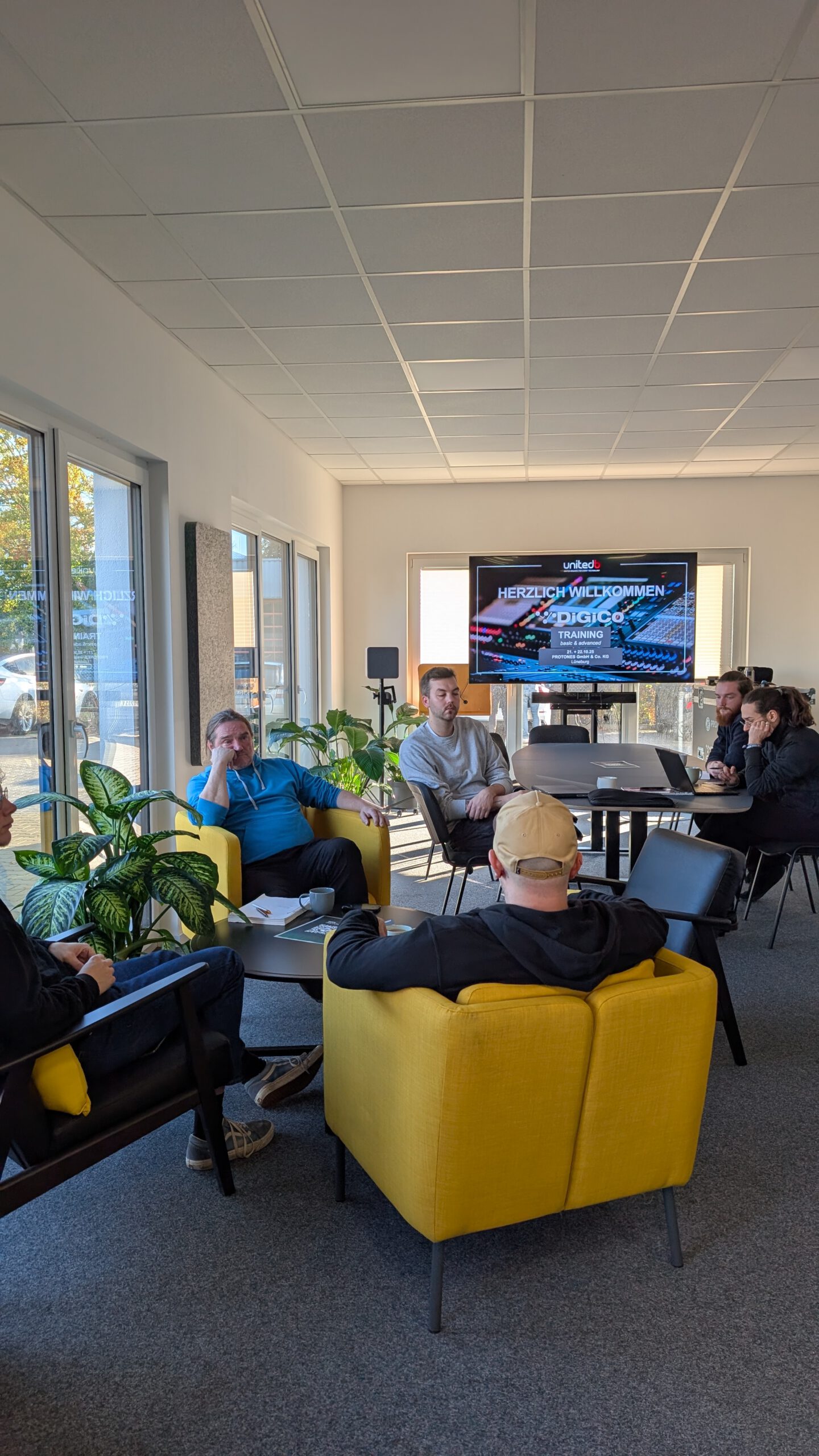 Teilnehmer sitzen in einem modernen Seminarraum bei der Begrüßung zum DiGiCo-Training, mit einem großen Bildschirm im Hintergrund, auf dem „Herzlich Willkommen zum DiGiCo Training“ zu lesen ist.