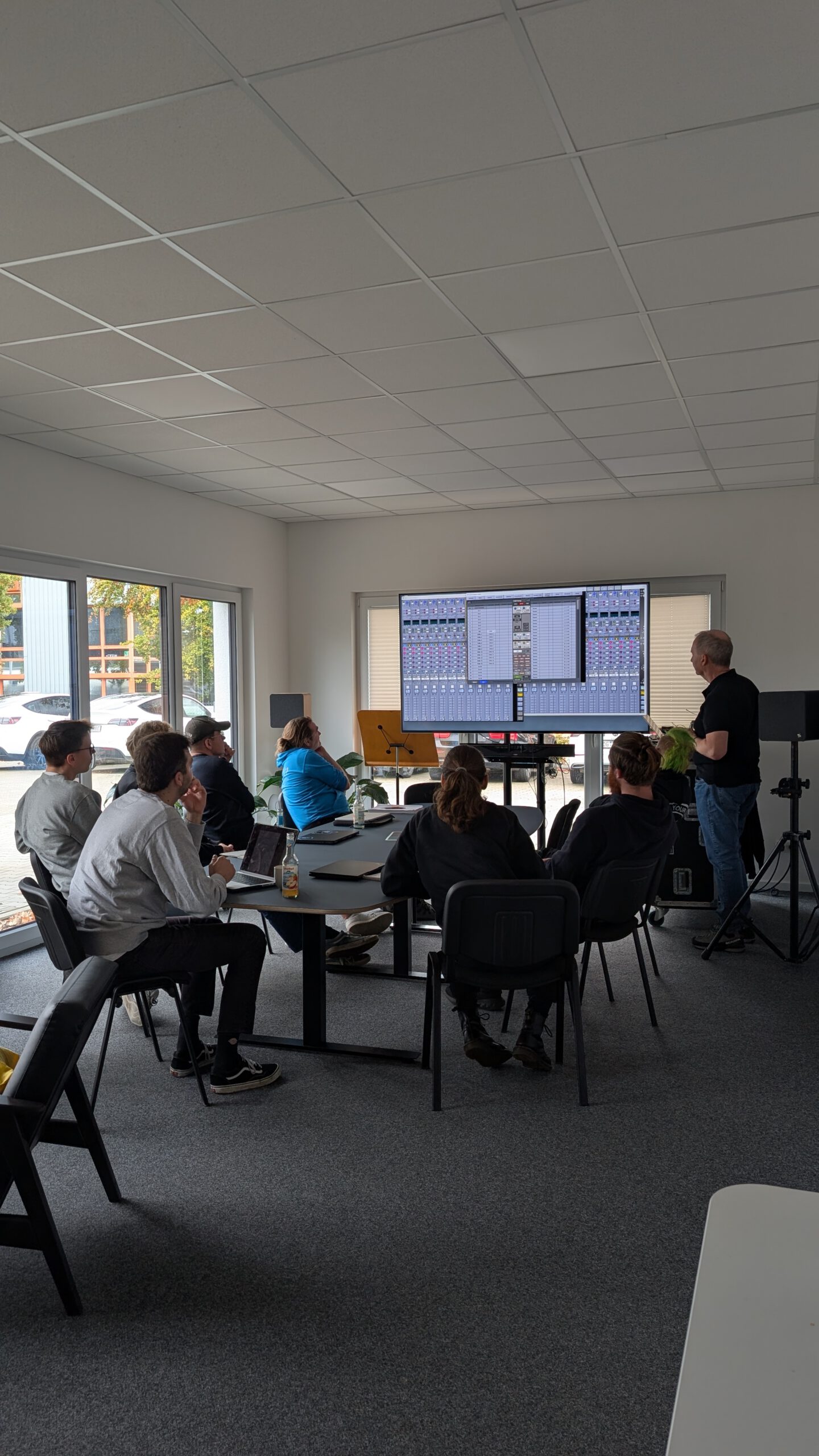 Teilnehmer einer Schulung sitzen in einem modernen Seminarraum und verfolgen eine Präsentation zur Bedienung einer Digitalmischpult-Software auf einem großen Bildschirm.